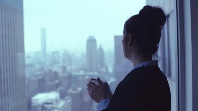 Eine Frau mit Tasse blickt aus einem Hochhausfenster auf eine verschneite Stadt
