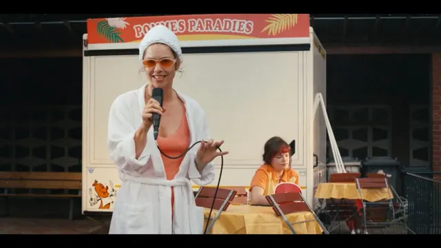 Eine Frau im Bademantel singt vor einem Imbisswagen mit der Aufschrift Pommes Paradies